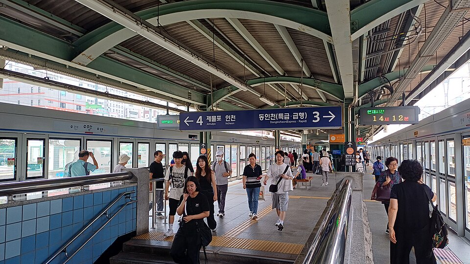 Covered train platform with passengers walking beneath a blue overhead sign showing platform numbers and destinations
