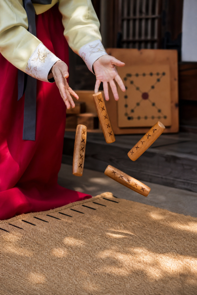 Four wooden yut sticks resting on a straw mat beside a Yutnori board