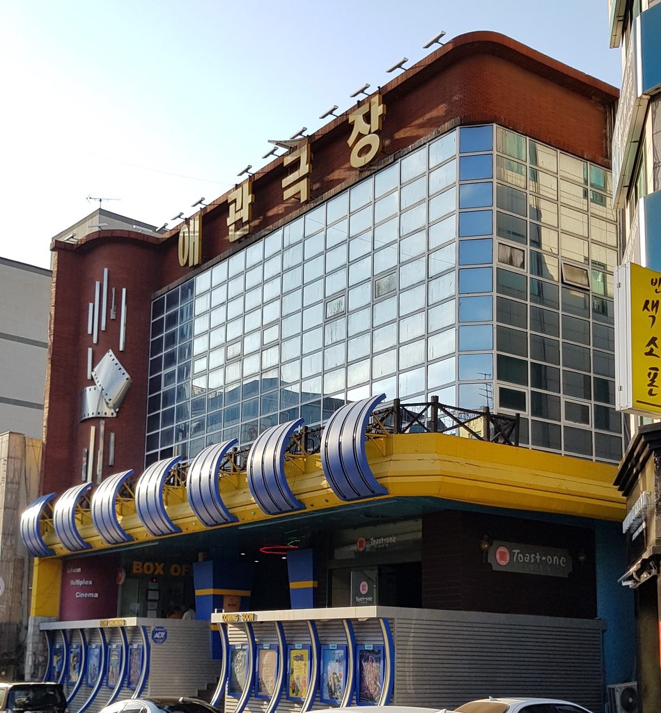 Curved brick and glass cinema building with large Korean lettering on the roof and a blue awning over the box office