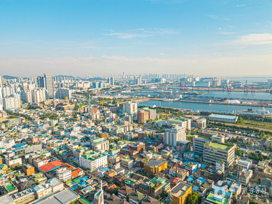 Aerial view of a dense low-rise neighborhood on a slope with container cranes and the harbor in the background