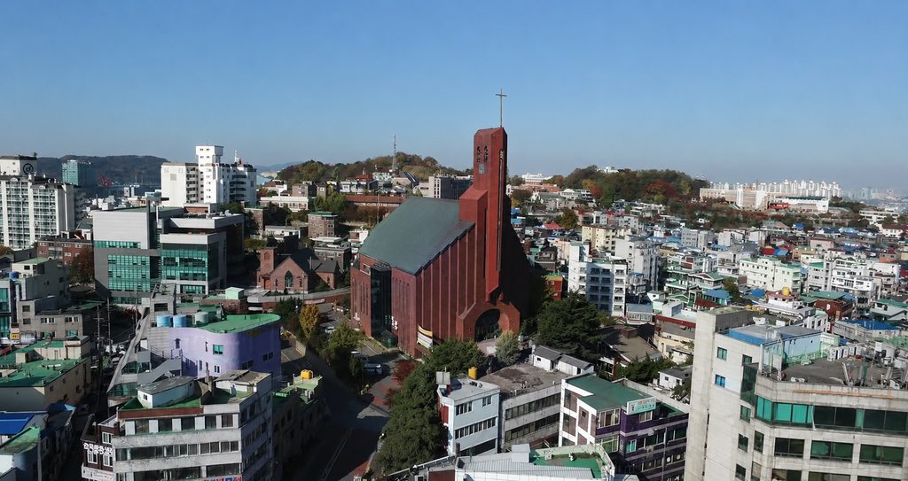 Tall red brick church with a green roof and slender bell tower rising above a dense hillside neighborhood with the harbor in the distance
