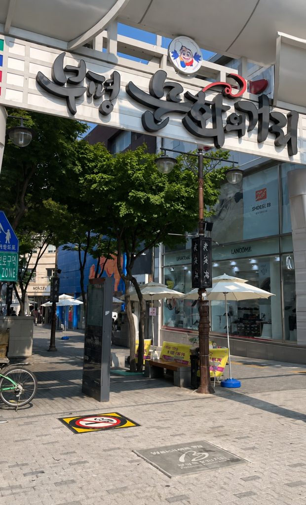 Pedestrian shopping street entrance with an arched sign, trees, and white parasols