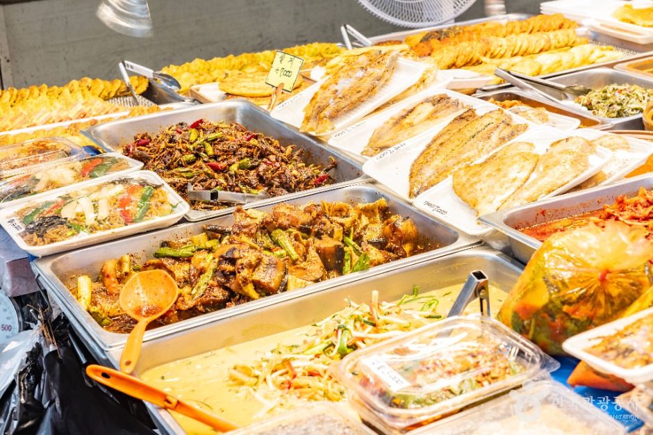 Metal trays of prepared side dishes and grilled fish displayed at a market stall