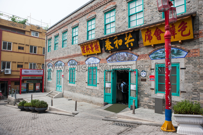 Two-story grey brick building with green window frames and painted wooden signboards