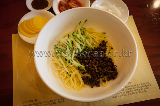 Bowl of yellow noodles topped with dark minced black bean sauce and shredded cucumber, with pickled radish on the side