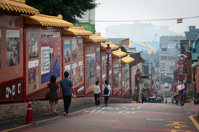 Long wall of painted story panels under small tiled eaves with people walking down a sloping street