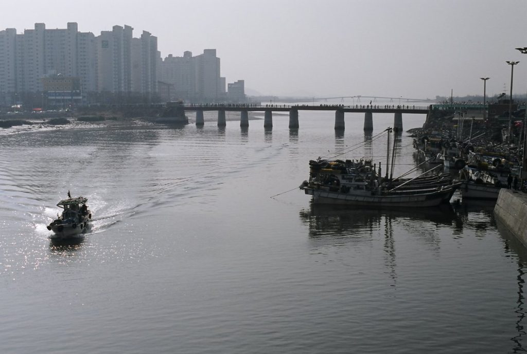 Long low footbridge crowded with people crossing a tidal channel, with a fishing boat passing beneath and apartment blocks behind